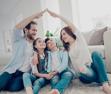Eine Familie sitzt eng nebeneinander im Wohnzimmer. Mutter und Vater sitzen jeweils außen und halten eine Hand hoch und verbinden diese in der Luft zu der Form eines Hausdachs.