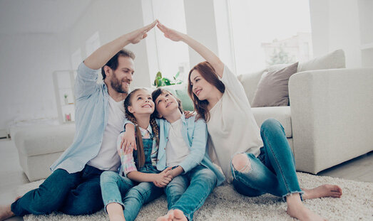 Eine Familie sitzt eng nebeneinander im Wohnzimmer. Mutter und Vater sitzen jeweils außen und halten eine Hand hoch und verbinden diese in der Luft zu der Form eines Hausdachs.