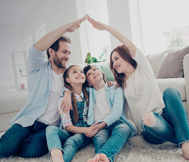 Eine Familie sitzt eng nebeneinander im Wohnzimmer. Mutter und Vater sitzen jeweils außen und halten eine Hand hoch und verbinden diese in der Luft zu der Form eines Hausdachs.