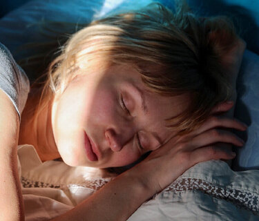 Eine Frau liegt schlafend im Bett. Die Sonnenstrahlen der hochfahrenden Rollos scheinen in den dunklem Raum auf ihr Gesicht.