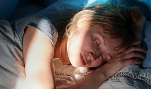 Eine Frau liegt schlafend im Bett. Die Sonnenstrahlen der hochfahrenden Rollos scheinen in den dunklem Raum auf ihr Gesicht.