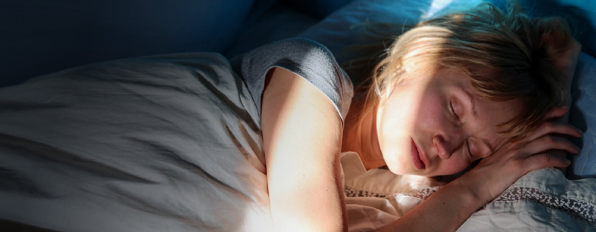 Eine Frau liegt schlafend im Bett. Die Sonnenstrahlen der hochfahrenden Rollos scheinen in den dunklem Raum auf ihr Gesicht.