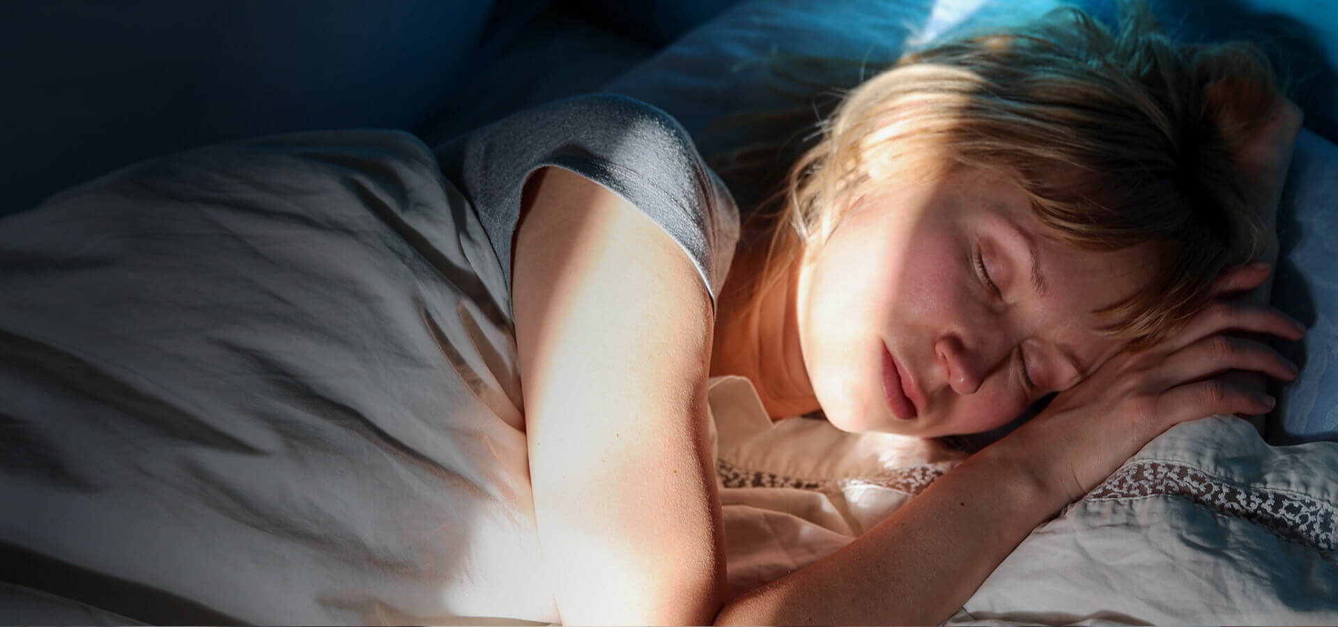 Eine Frau liegt schlafend im Bett. Die Sonnenstrahlen der hochfahrenden Rollos scheinen in den dunklem Raum auf ihr Gesicht.