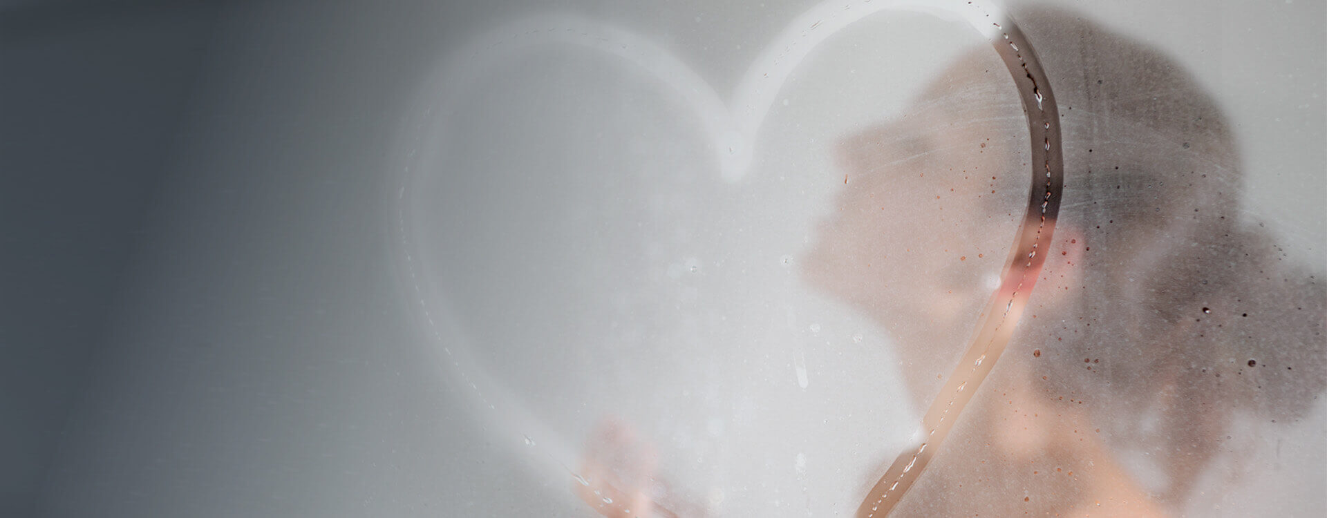 Eine Frau steht in der Dusche. In der beschlagenen Duschwand ist ein Herz gemalt.  