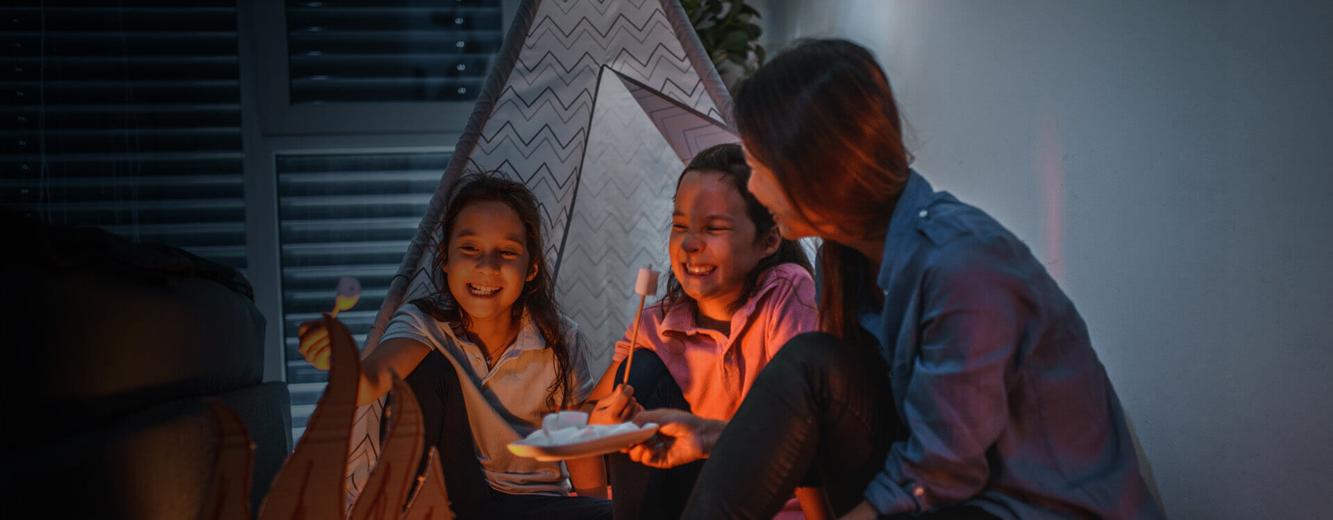 Eine Familie sitzt im dunklen Kinderzimmer in einem Kinderzelt, die Rollos sind heruntergefahren.