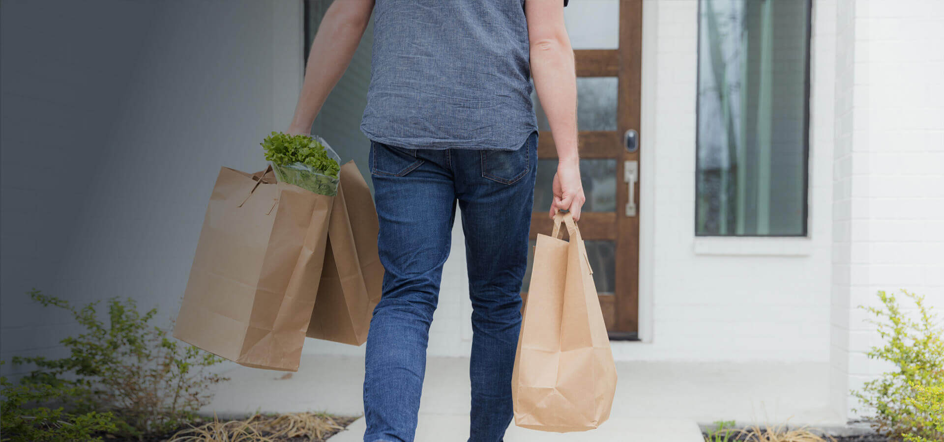 Eine Frau läuft auf ihre Haustür zu, in beiden Händen hält sie eine Einkaufstüte.