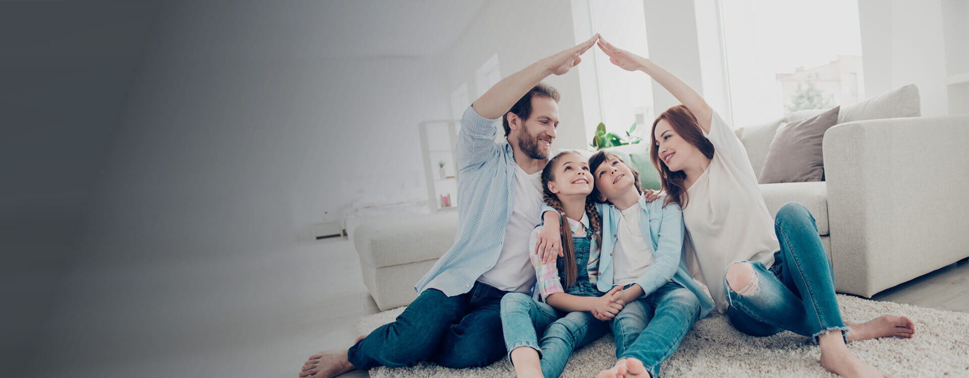 Eine Familie sitzt eng nebeneinander im Wohnzimmer. Mutter und Vater sitzen jeweils außen und halten eine Hand hoch und verbinden diese in der Luft zu der Form eines Hausdachs.