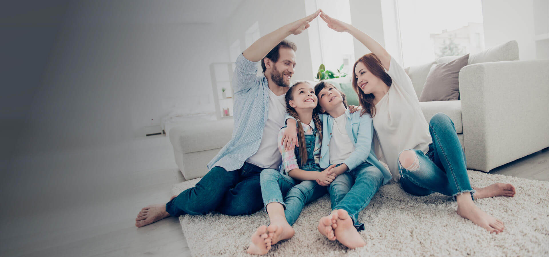 Eine Familie sitzt eng nebeneinander im Wohnzimmer. Mutter und Vater sitzen jeweils außen und halten eine Hand hoch und verbinden diese in der Luft zu der Form eines Hausdachs.